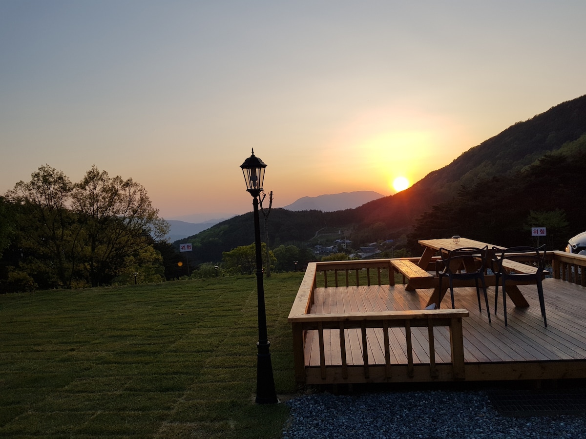 A wooden deck is seen extending into a serene landscape, with a sunset casting warm hues over the distant mountains. A lamp post stands near the deck, enhancing the tranquil setting, while freshly mowed grass creates an inviting outdoor space.