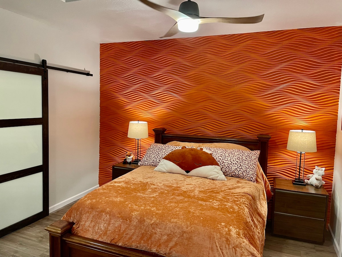 A cozy sleeping area features a warm orange accent wall with a textured wave design. A spacious bed is dressed in an orange blanket, complemented by decorative pillows. Two bedside tables hold matching lamps, providing soft lighting, while a sliding door adds a contemporary touch.