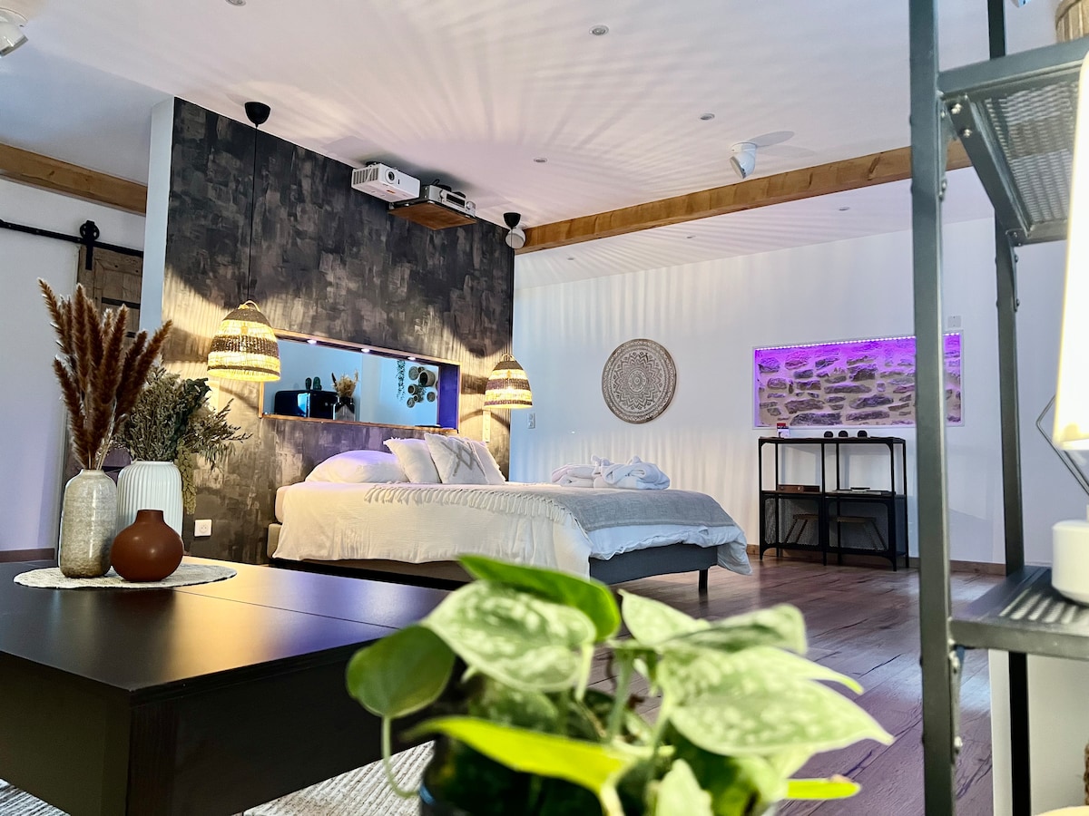 A spacious room is presented with neutral tones and wooden accents. Soft lighting from two modern pendant lamps creates an inviting ambiance. A large bed is positioned against a textured feature wall, and a small seating area is visible, complemented by decorative plants and an accent table.