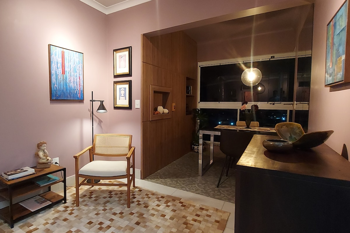 A cozy corner in the living area is highlighted by a rattan chair positioned beside a small bookshelf. The walls are adorned with framed artwork, and the adjacent space features a modern dining area with large windows allowing for ample natural light.