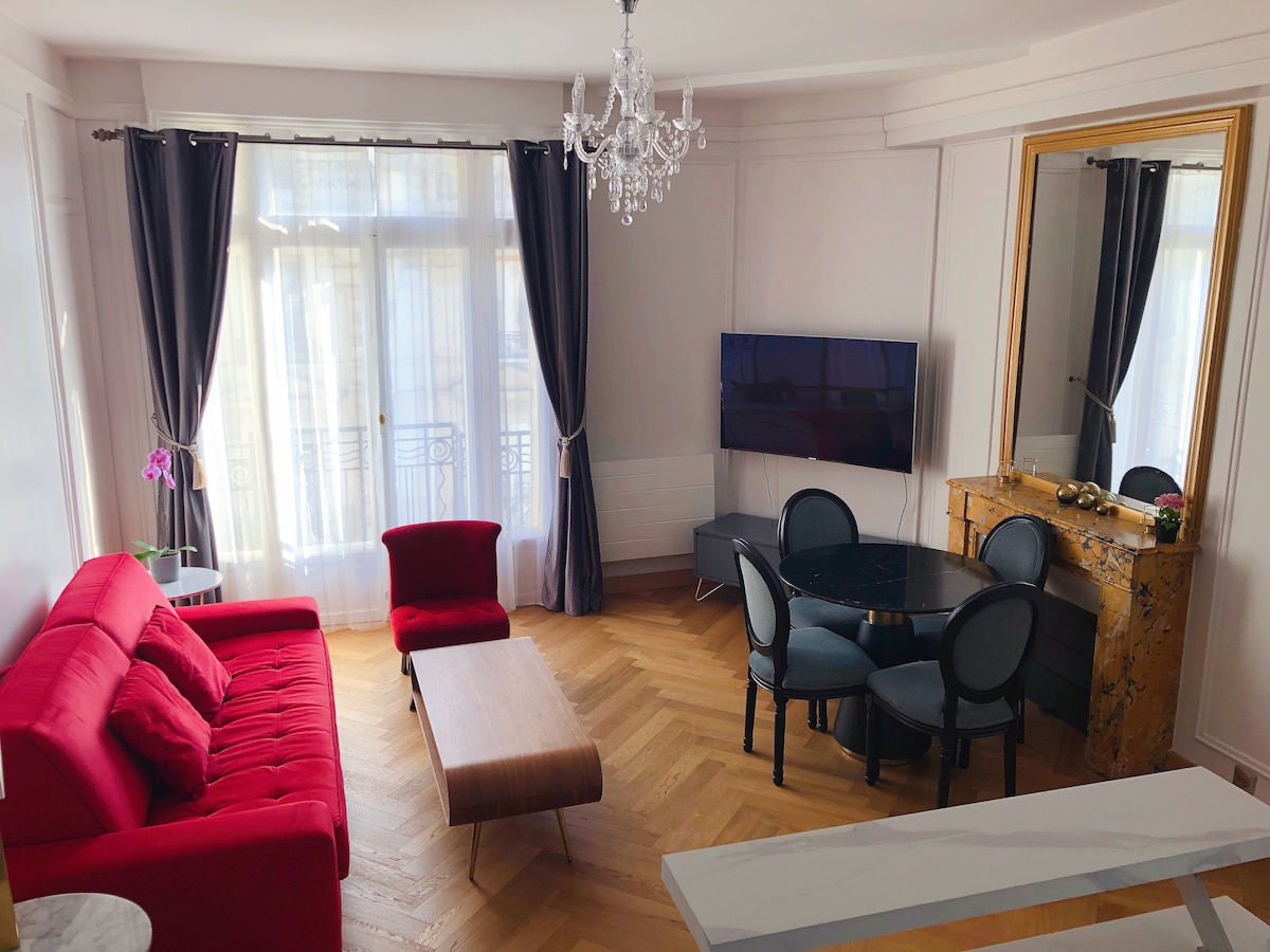 A spacious living area features a comfortable red sofa and a sleek wooden coffee table. A round dining table with black chairs is positioned nearby. Natural light fills the room through large windows, which are draped with sheer curtains. A decorative mirror and a chandelier add elegance to the space.