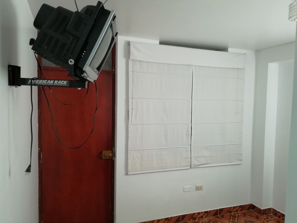 A wall-mounted television is positioned above a door, which features a wooden finish. A window with white blinds allows natural light to enter the room, while the floor is covered with decorative tiles, adding visual interest.