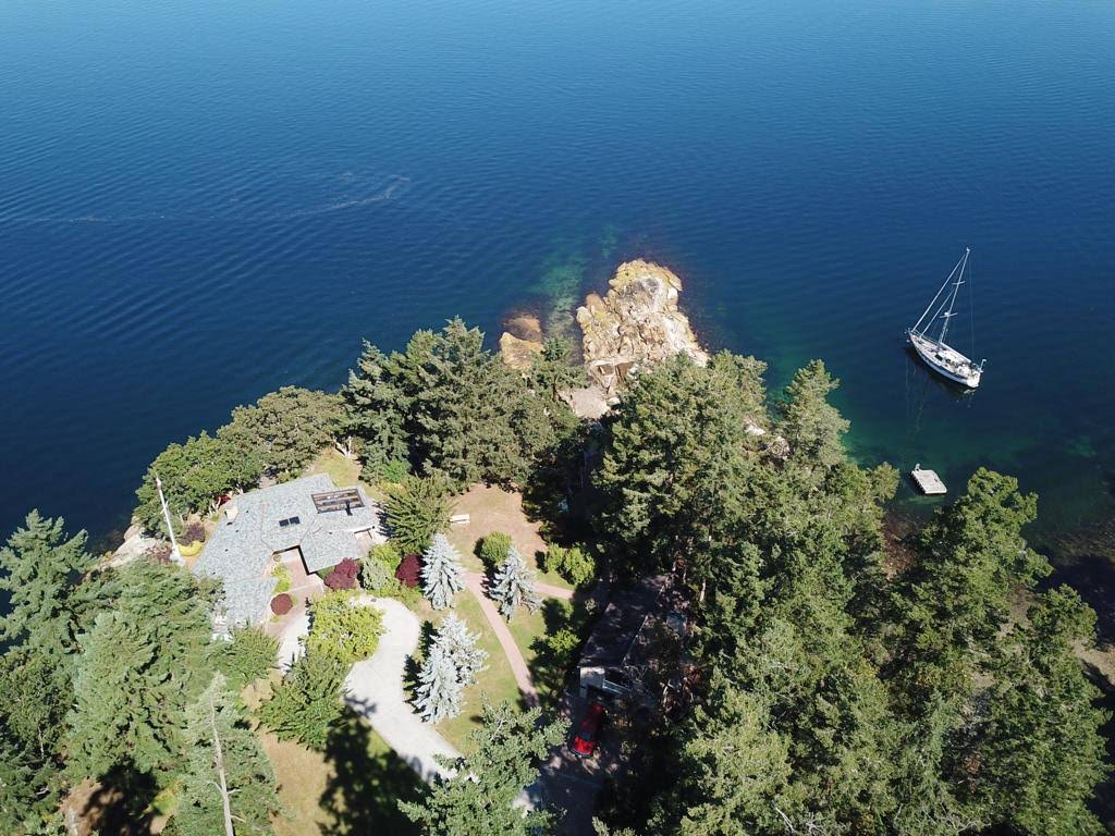 An aerial view captures the estate set against a vibrant blue ocean. Surrounding greenery includes trees and well-maintained gardens, while a sailboat is anchored nearby, accentuating the property's waterfront location.