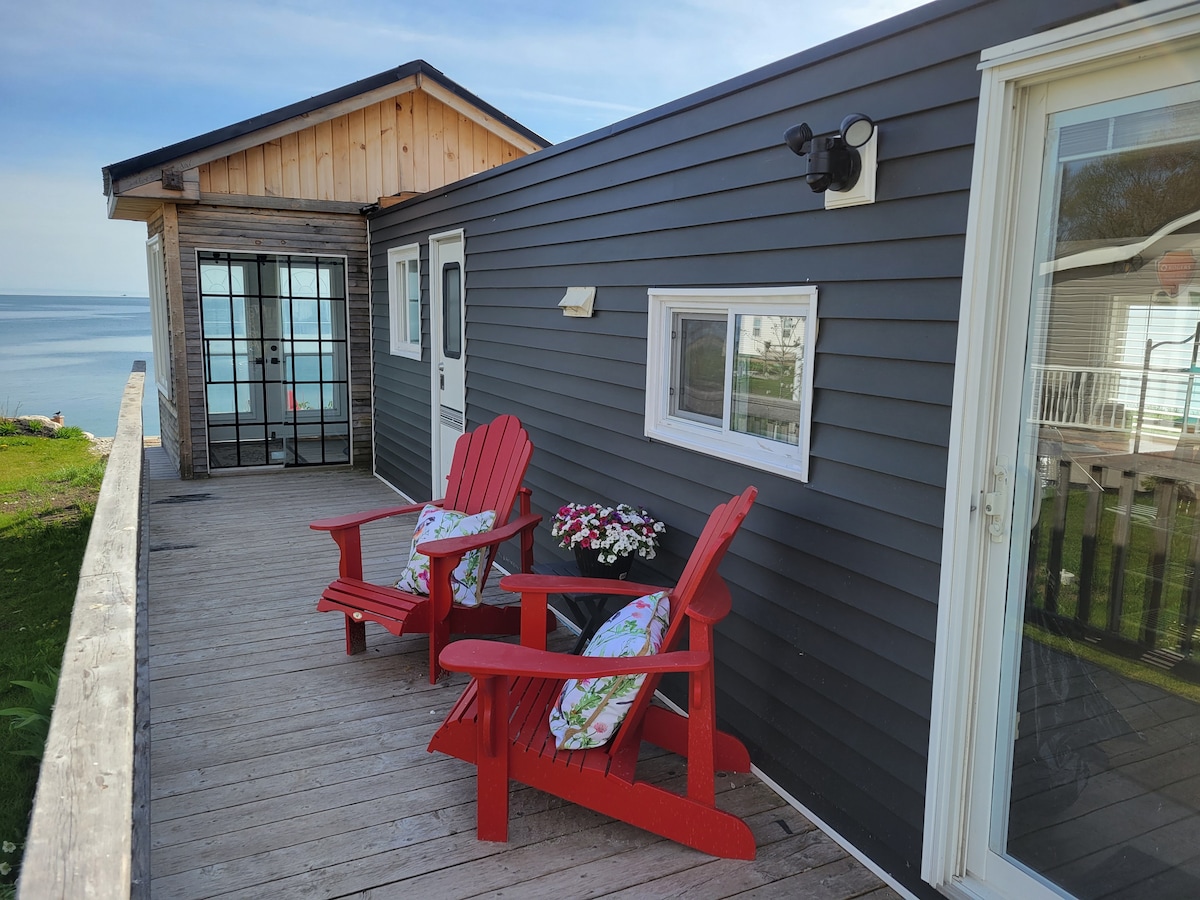 A wooden deck features two red Adirondack chairs, each adorned with floral cushions. A potted plant with bright flowers is situated between the chairs. The backdrop includes a view of the lake and a neighboring building with wood accents.