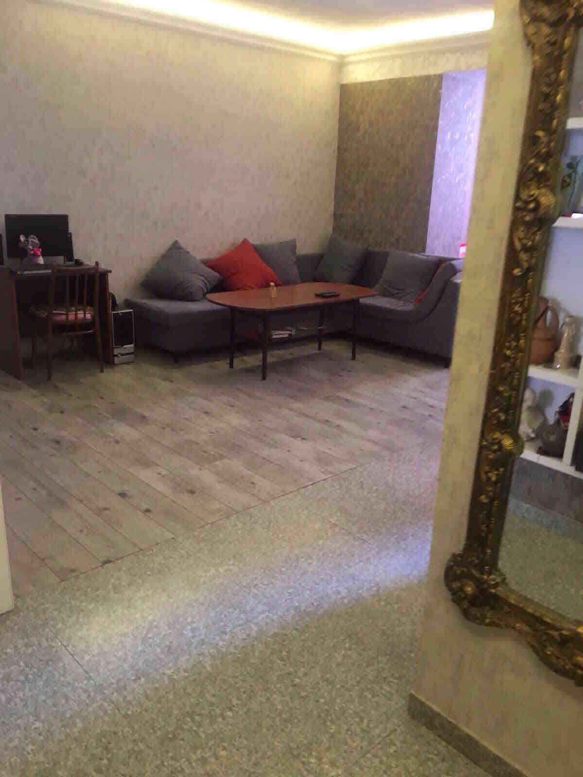 The living area features a grey sectional sofa adorned with decorative pillows and a low coffee table. A small desk is positioned against the wall, accompanied by a computer. The flooring is light wood, and soft lighting enhances the overall ambiance.
