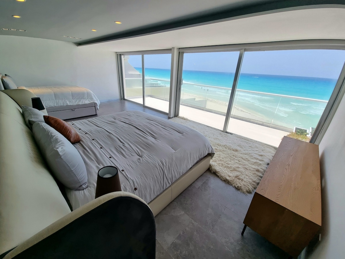 A spacious bedroom showcases large windows that offer unobstructed ocean views. Two beds are positioned within the room, complemented by a light-colored rug beneath them. A wooden dresser stands against one wall, enhancing the minimalist design.