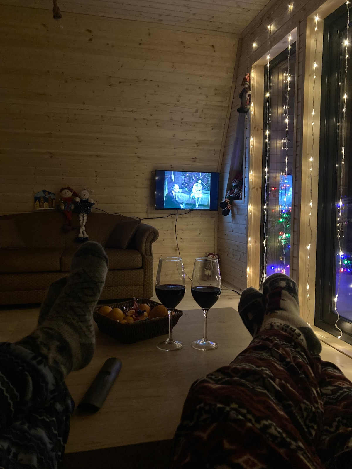 A cozy living area is shown with a wooden interior. A sofa is positioned in the background, along with a television displaying a performance. Two glasses of wine rest on a table, accompanied by a bowl of fruit. Soft lighting from fairy lights enhances the ambiance.
