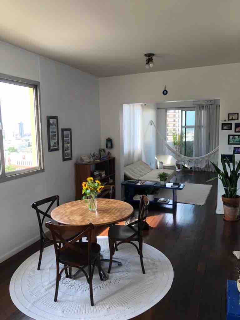 A bright living and dining area features a round wooden table surrounded by four chairs. A light-filled corner showcases a white hammock and a soft seating arrangement, complemented by potted plants. Large windows provide a view of the city skyline, enhancing the spacious ambiance.