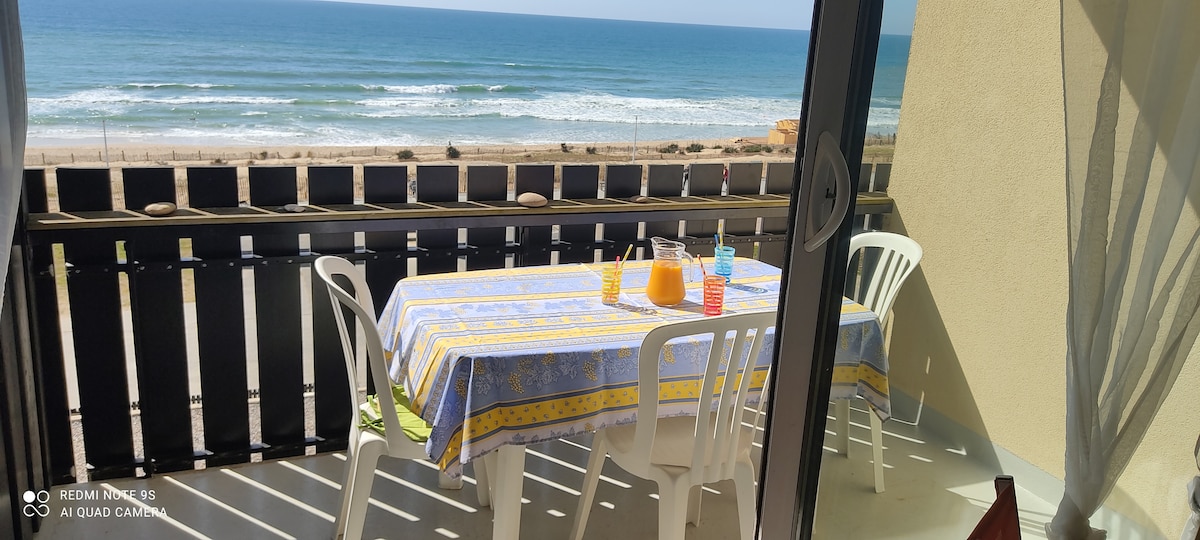 A spacious balcony is presented with a table set for four, featuring a vibrant tablecloth. Refreshing beverages, including orange juice, are displayed on the table. The expansive ocean view is visible beyond the railing, with gentle waves meeting the sandy shore.