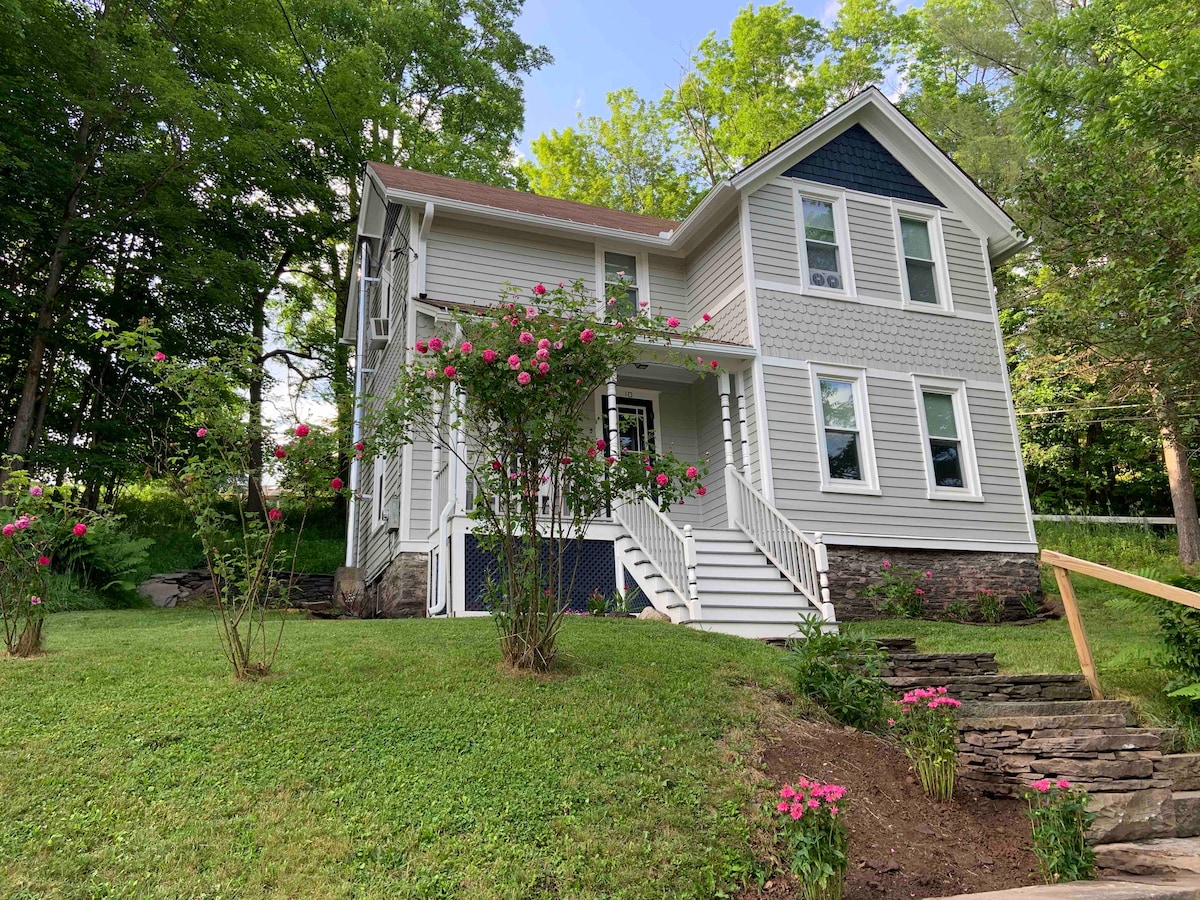 A three-story house is set amidst lush greenery, framed by flowering shrubs. The front entrance features a set of white stairs leading to an inviting porch. Large windows allow natural light to fill the interior, while a mix of blue and gray siding adds character.