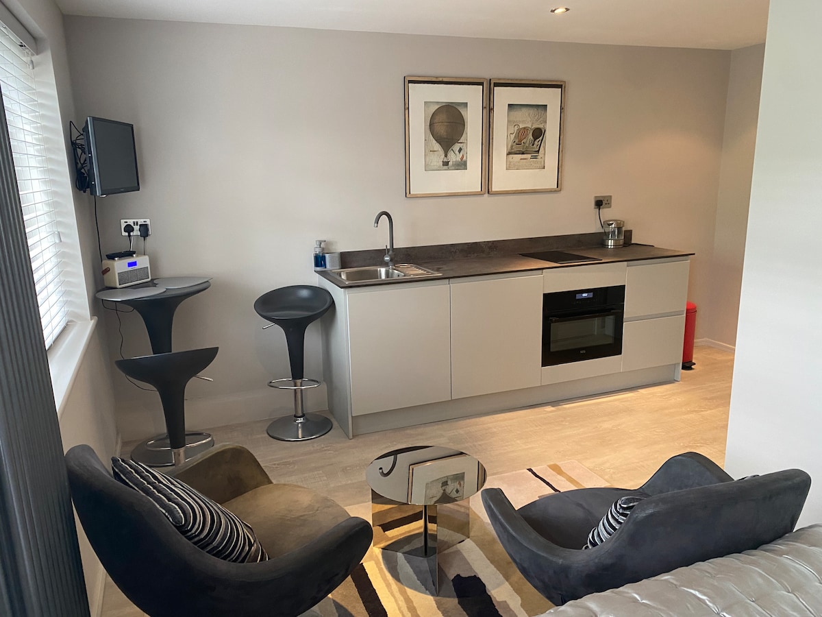 A modern kitchen area is visible, featuring sleek cabinetry and stainless steel appliances. A small dining table with two high stools is positioned beside a cozy seating area comprised of two plush chairs and a contemporary coffee table. Artwork adorns the walls, adding a touch of character.