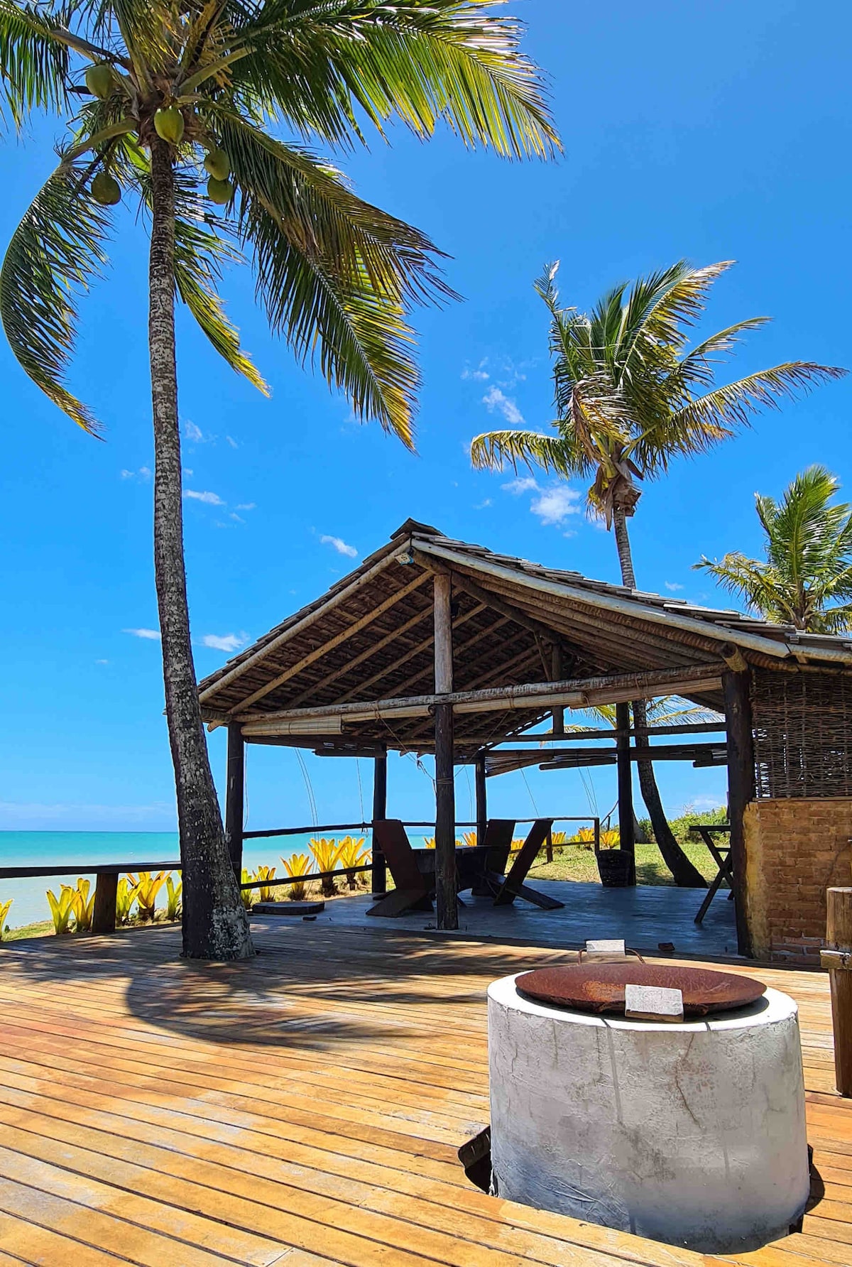 A shaded outdoor area features a traditional structure with a thatched roof, supported by wooden beams. The space is surrounded by palm trees, with a wooden deck offering a view of the calm sea. A circular fire pit stands on the deck, inviting relaxation.