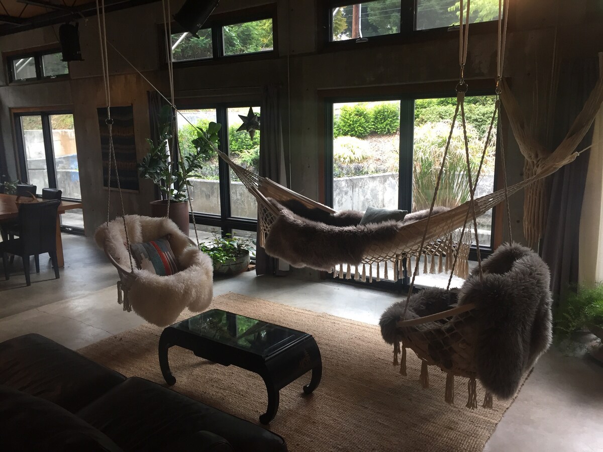 Two hanging chairs are suspended from the ceiling, each draped with soft, cozy blankets. A large area rug covers the concrete floor, and natural light floods the space through expansive windows, illuminating surrounding greenery visible outside.