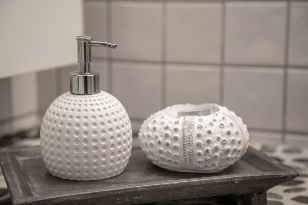 A stylish bathroom countertop is adorned with a pump dispenser and a matching decorative holder. Both items feature textured surfaces resembling sea urchins, presented on a sleek tray. The muted background includes square grey tiles that contribute to a clean and modern aesthetic.
