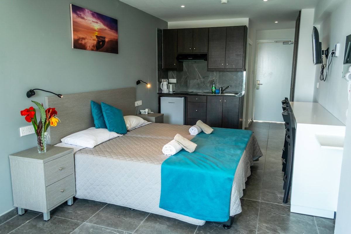A modern studio space is highlighted by a spacious bed adorned with turquoise and white linens. A kitchenette is visible in the background, equipped with essential appliances. Natural light enters through a window, complementing the neutral grey walls. A small vase with flowers adds a touch of color.