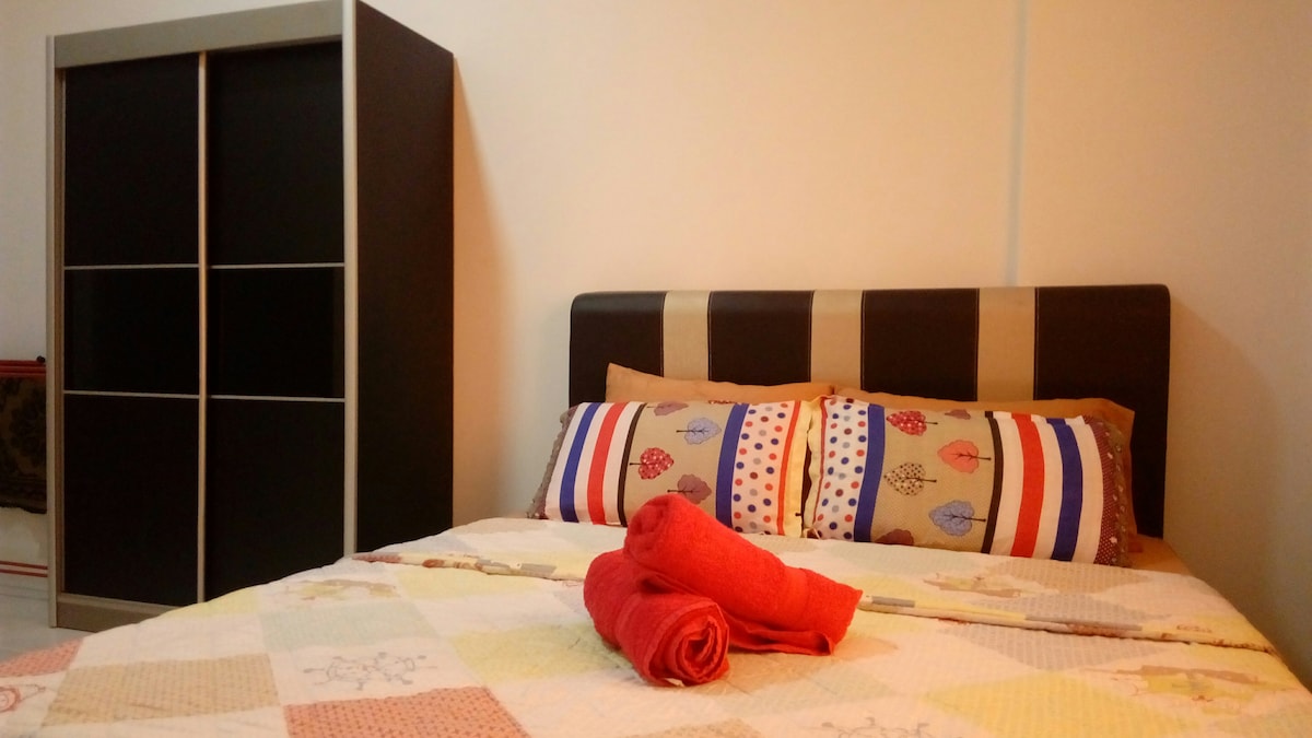 A well-appointed bedroom is showcased, featuring a spacious bed adorned with patterned pillows and a colorful quilt. Neatly arranged red towels are placed at the foot of the bed. A sleek wardrobe stands against the wall, providing ample storage space.