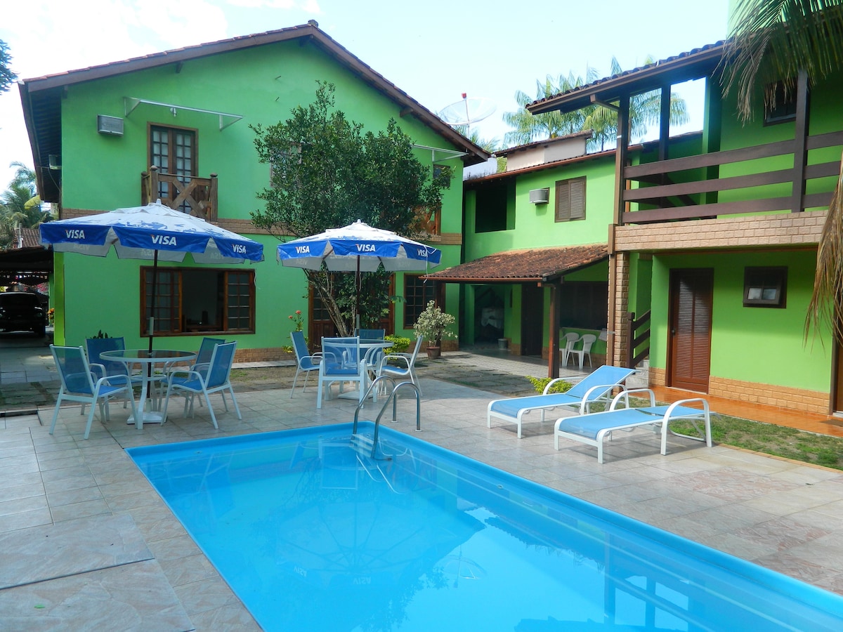 A tranquil outdoor area features a refreshing swimming pool surrounded by lounge chairs. Umbrellas provide shade over tables nearby. The vibrant green exterior of the buildings creates a lively backdrop, with palm trees adding to the inviting setting.