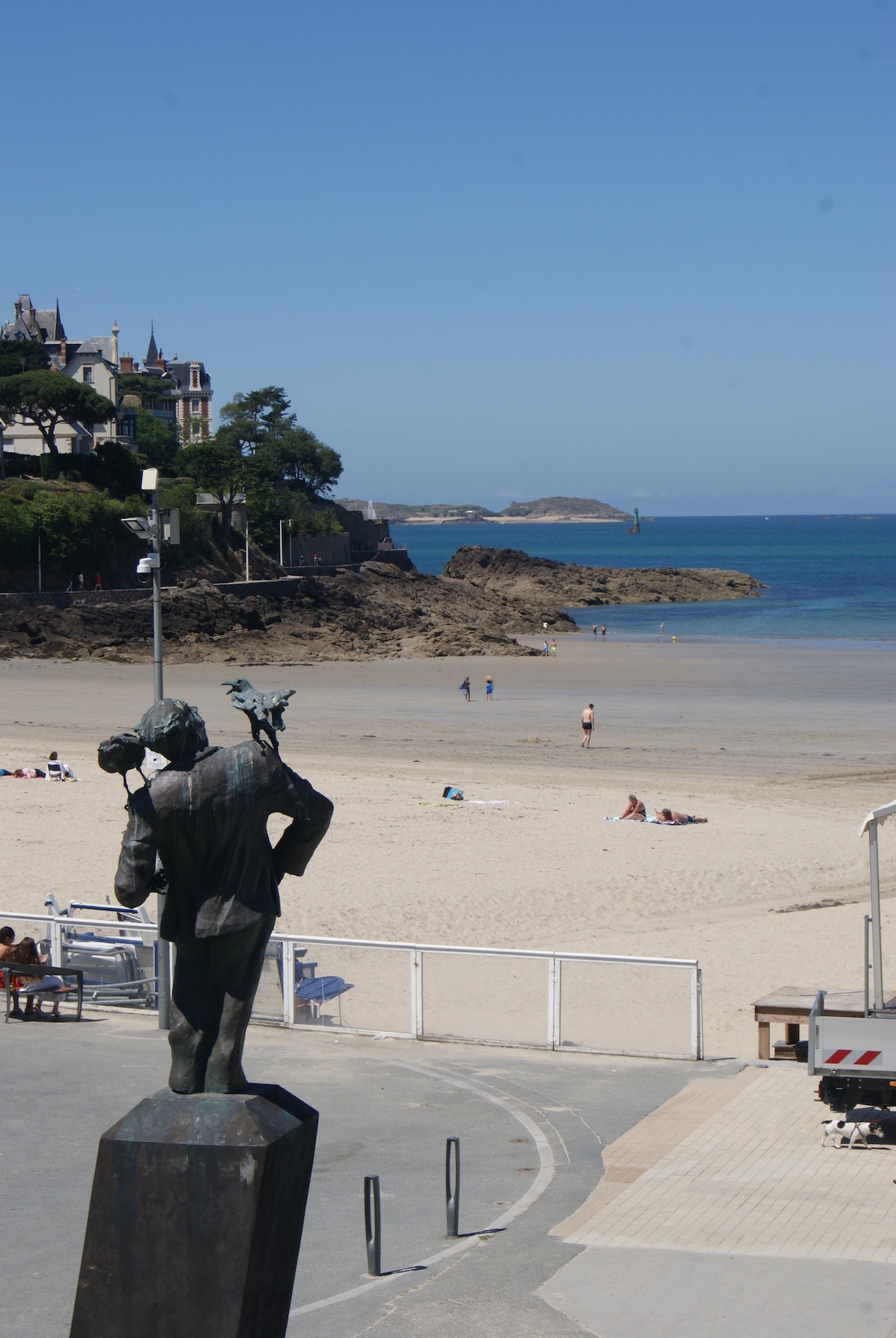 A bronze statue stands prominently overlooking a sandy beach with a calm sea in the background. Sunbathers are visible on the beach, while rocky formations can be seen off the shore. Coastal buildings frame the scene, contributing to the tranquil atmosphere.
