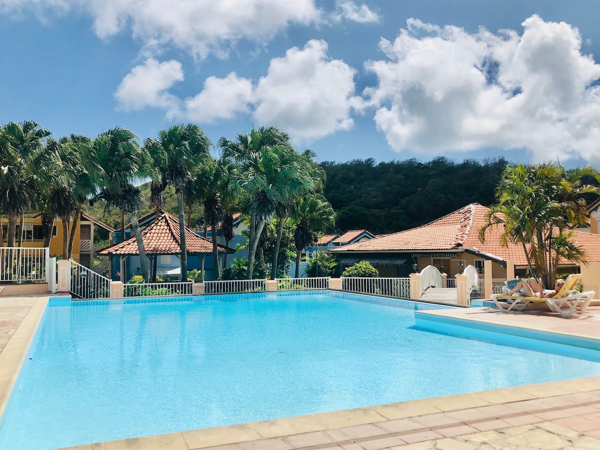 A large, inviting swimming pool is surrounded by lush palm trees and tropical landscaping. The clear blue water reflects the sky, while shaded lounge chairs are positioned on the pool deck. Nearby, low-rise buildings with terracotta roofs complement the serene ambiance.