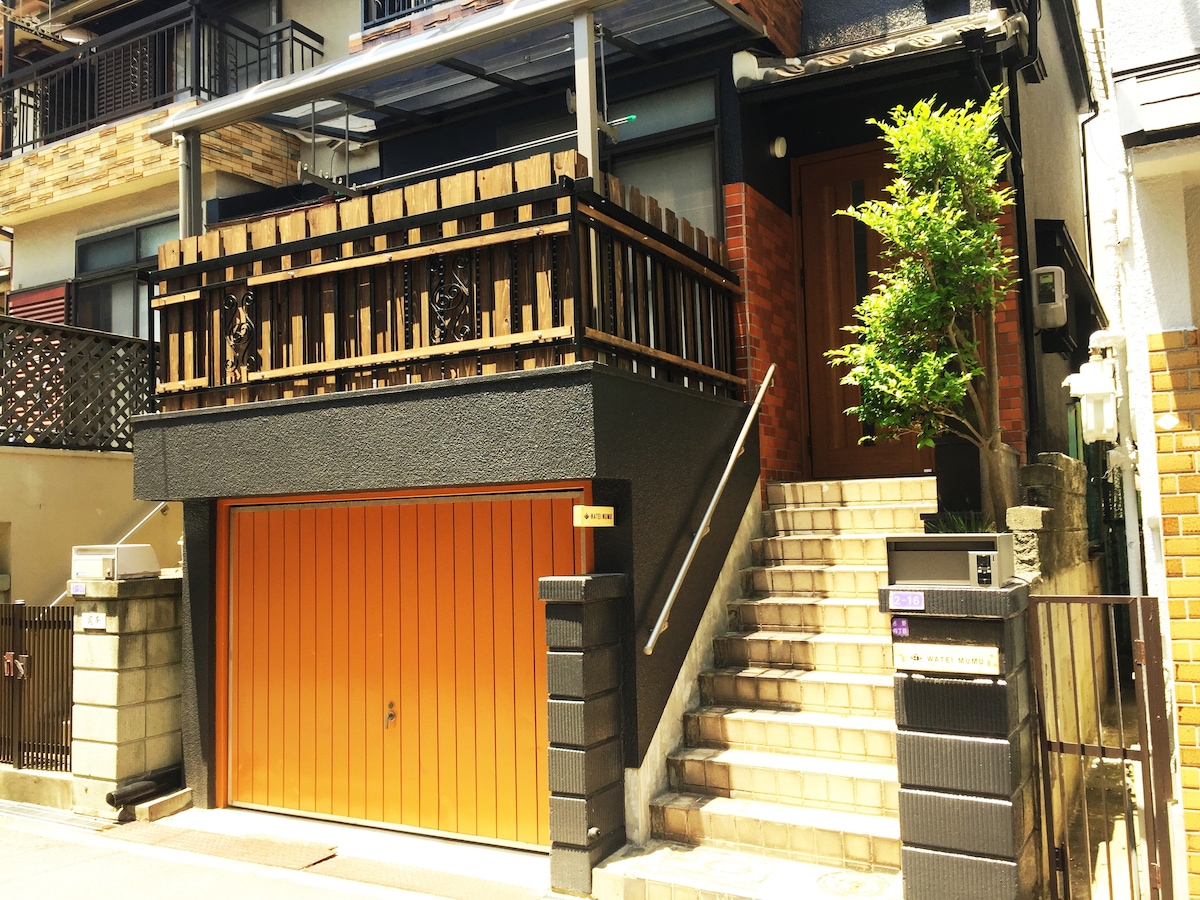 A three-story building is pictured, featuring a garage with a vibrant orange door. Stairs lead to the entrance, enhanced by a small tree near the door. The exterior displays a blend of natural wood and stone elements, presenting a welcoming façade.