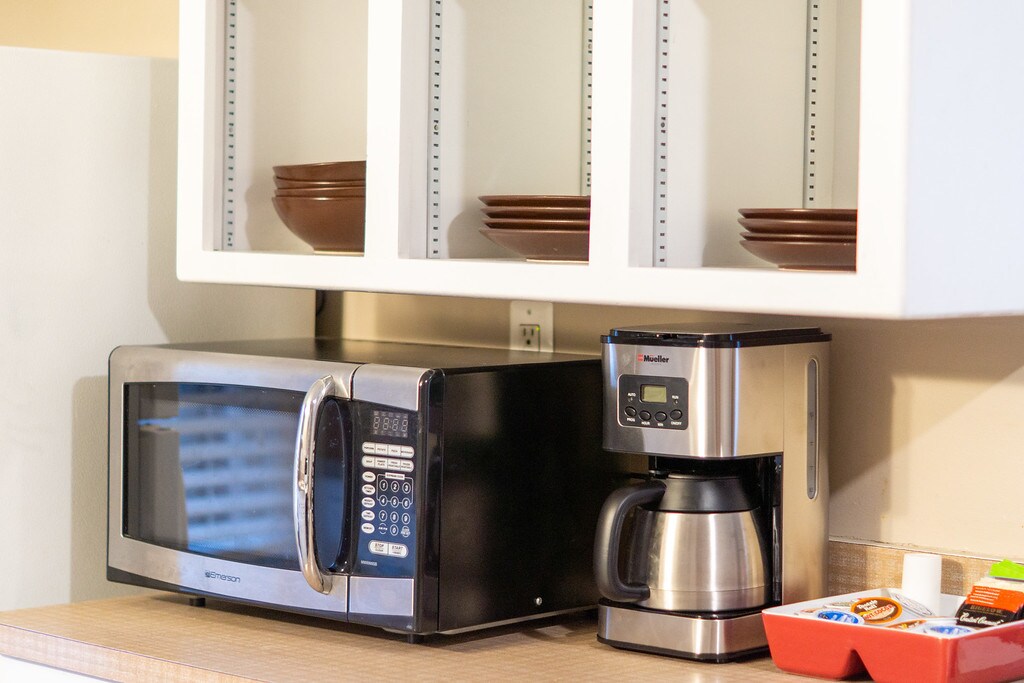 A kitchenette is equipped with a microwave and a coffee maker on a countertop. Above the appliances, open shelving displays brown plates, while a tray holds various snack items, creating a functional and easily accessible space for meal preparation.