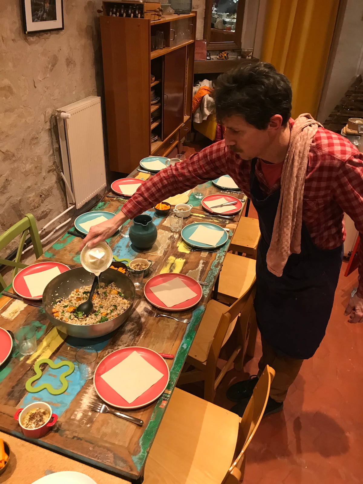 A dining table is set with colorful plates and napkins, surrounded by wooden chairs. A person is pouring a liquid into a large cooking pot placed at the center of the table. The backdrop includes rustic stone walls and wooden cabinets, adding a homely touch to the space.