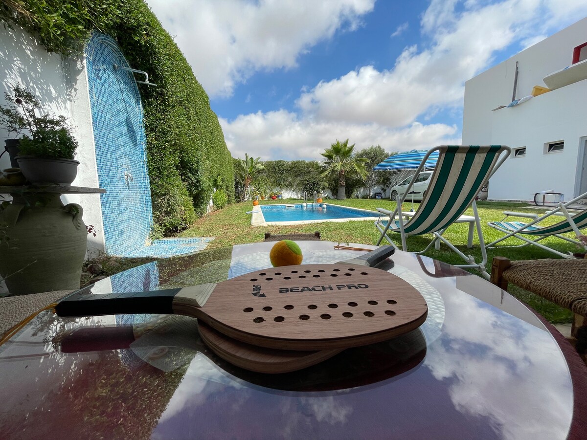 A wooden paddle rests on a glass table, while a lemon sits nearby. In the background, a swimming pool is visible, surrounded by greenery and sun loungers. The sky features a mix of clouds and blue, creating a serene atmosphere.