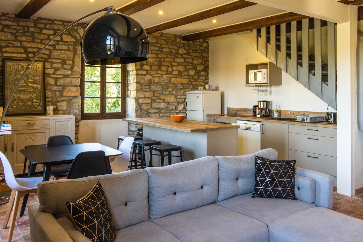 A spacious living area features a comfortable gray couch accented with decorative pillows. A modern dining table is accompanied by white chairs, while a kitchen area with sleek cabinetry and appliances is visible in the background. Natural light filters in through multiple windows, illuminating the rustic stone walls.