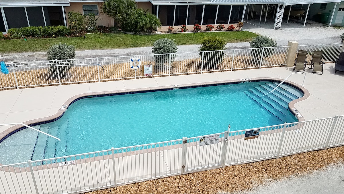 A private pool area is enclosed by a white fence, showcasing clear turquoise water. Steps lead into the pool on one side, while lounge chairs sit nearby. The surrounding landscape includes carefully maintained greenery and colorful plantings, contributing to a relaxing outdoor space.