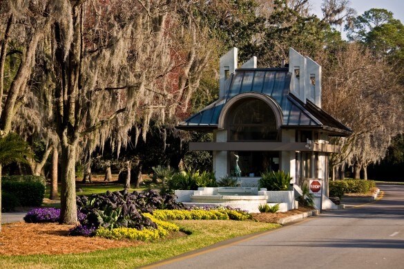 A welcoming entrance to the gated Shipyard Plantation is shown, featuring a modern gatehouse complemented by lush landscaping. Green grass and vibrant flower beds featuring purple and yellow blooms frame the entrance. Spanish moss hangs from tree branches in the background, enhancing the serene atmosphere.