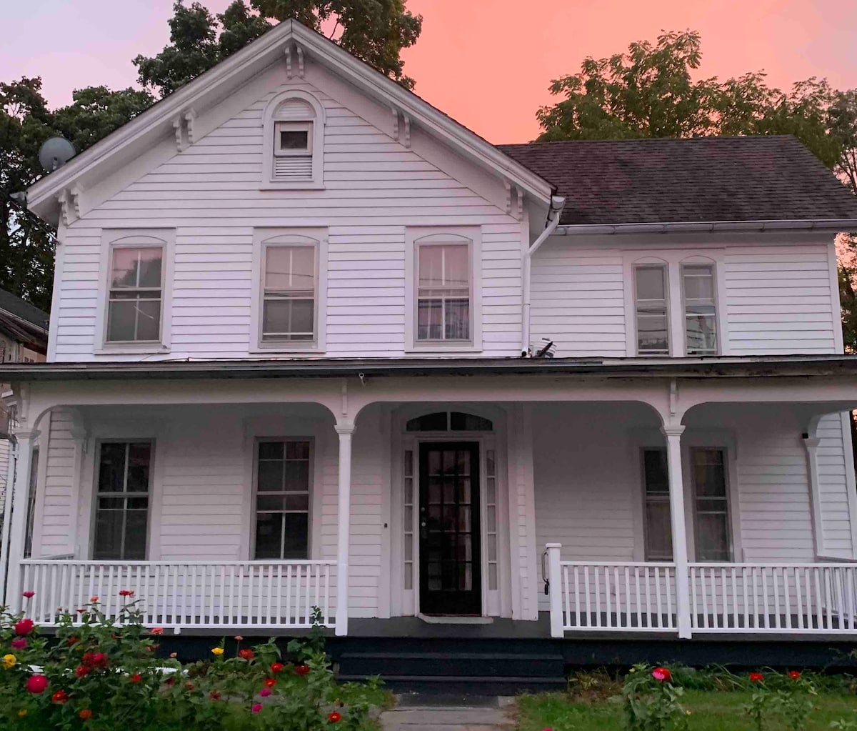 A charming two-story white house is framed by a pastel sunset sky. The front porch features decorative columns and a black door, with multiple windows allowing for natural light. A flower bed with blooming flowers adds a touch of color to the foreground.