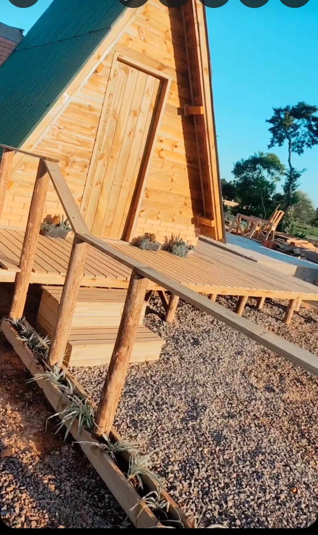 The entrance to a wooden cabin is depicted, featuring a ramp leading up to the door. Surrounding the ramp, small plants are visible along the edge, and the cabin's sloped roof and wooden exterior add a rustic charm. The clear sky enhances the inviting atmosphere.