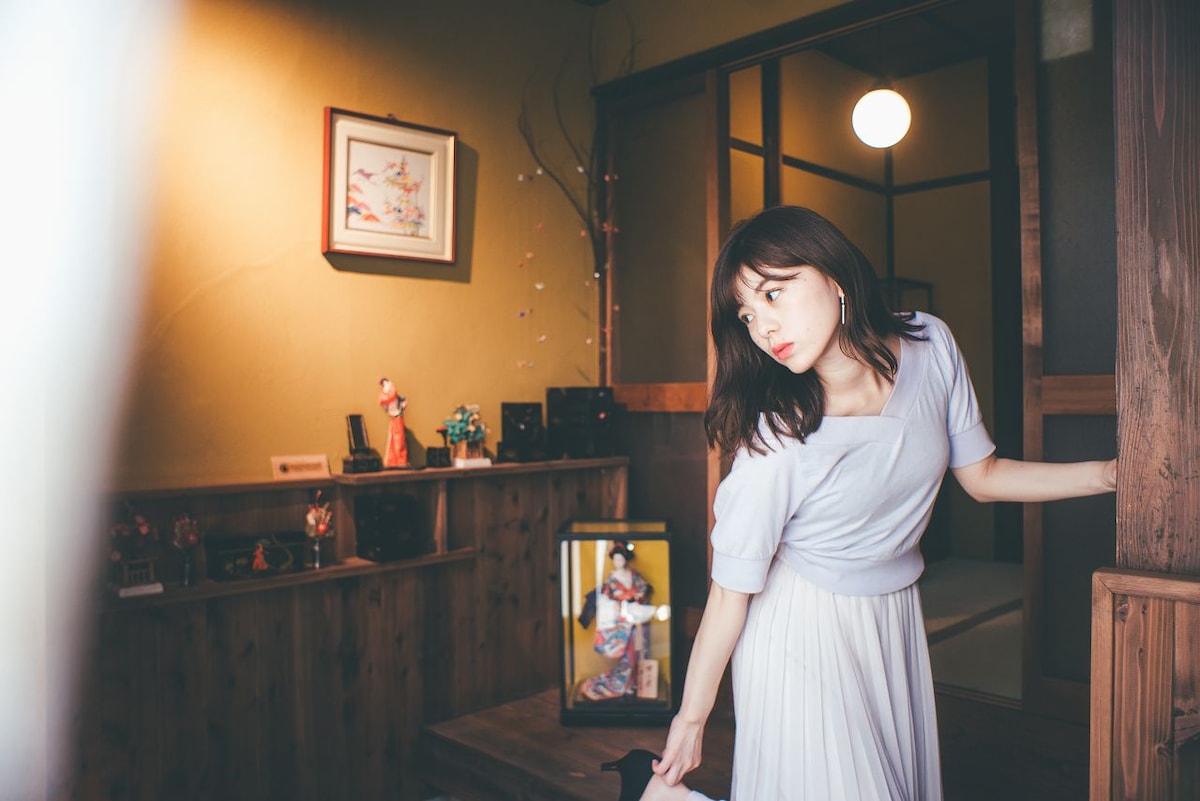 A woman stands in the doorway of a traditional Japanese room, illuminated by warm light. A decorative shelf featuring various figurines and artwork is visible in the background, complemented by a floor mirror reflecting the inviting interior.