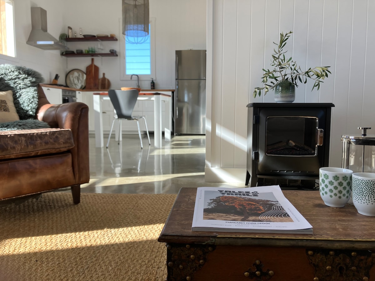A welcomely arranged interior features a cozy living space with a leather sofa and a coffee table displaying a magazine and two cups. The kitchen, visible in the background, includes modern appliances and is complemented by wooden shelves adorned with decorative items.