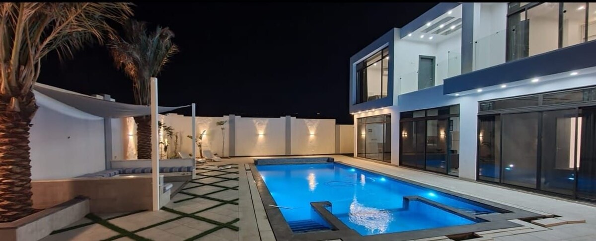 A modern outdoor space is shown at night, featuring a well-lit swimming pool that reflects soft blue tones. Surrounding palm trees add greenery, while the bright facade of the house is lined with large glass doors. Lounge chairs are positioned for relaxation.