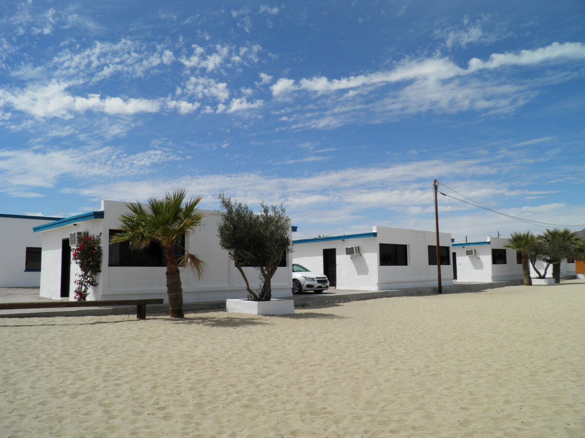 The image displays a row of white buildings with blue accents against a clear sky. Palm trees and shrubs are positioned in front, and a sandy area leads up to the structures. A vehicle is parked nearby, enhancing the beachside setting.