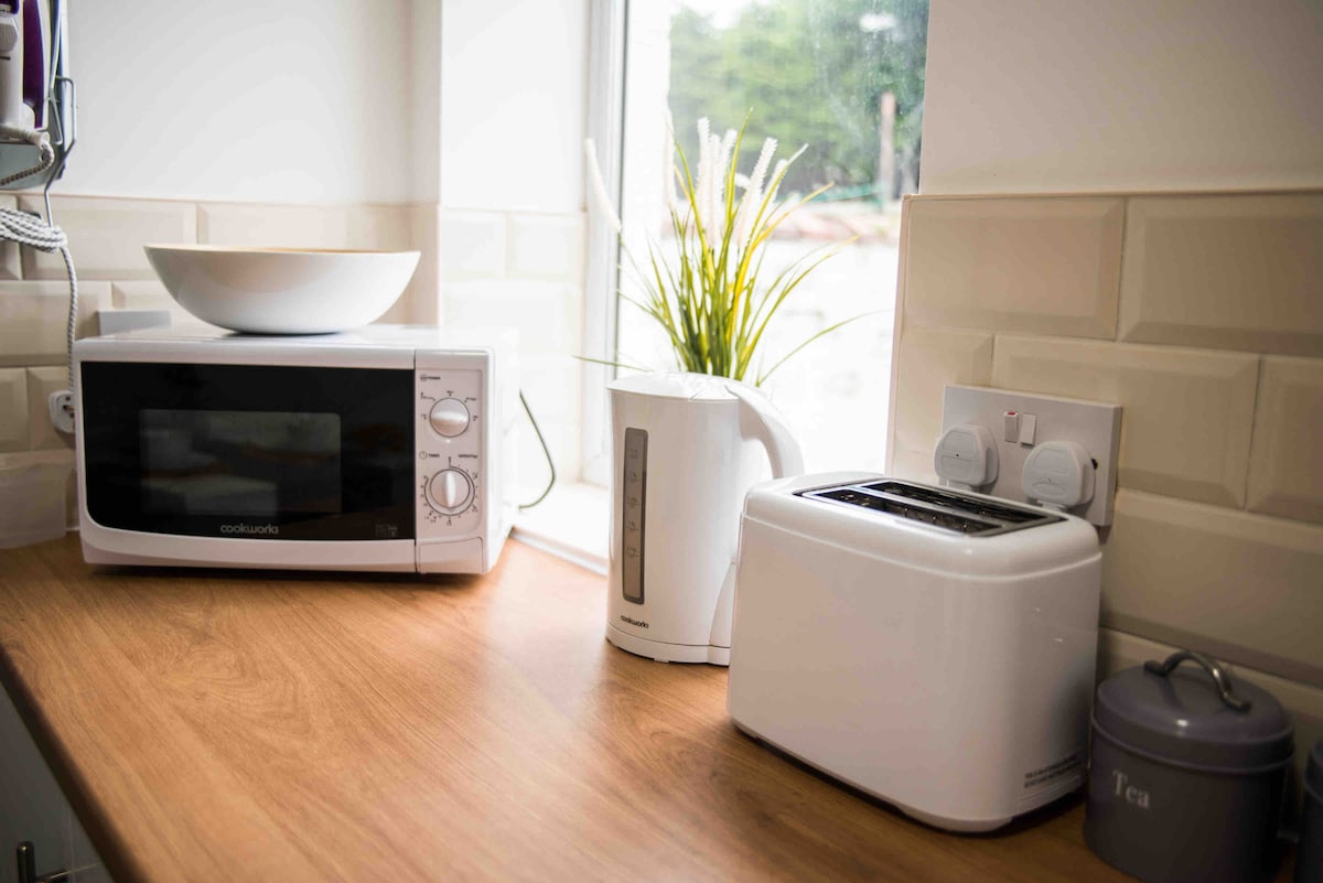 A kitchen countertop is presented with a microwave, kettle, and toaster, arranged neatly. A bowl is placed on top of the microwave. Soft natural light enters through a nearby window, illuminating a potted plant positioned against the wall.