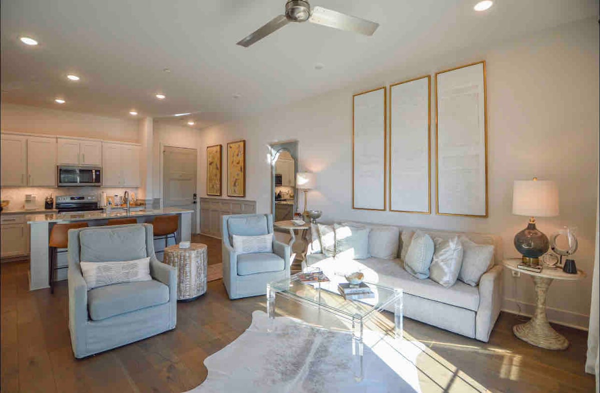 The open-concept living area features a light-colored sofa and two armchairs, complemented by a glass coffee table. A ceiling fan hangs above, while kitchen cabinetry is visible in the background. Natural light fills the space through recessed lighting and nearby windows.