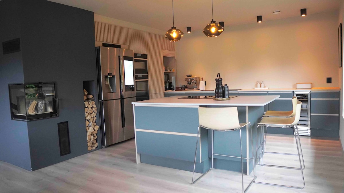 A modern kitchen is featured, showcasing a central island with bar stools and sleek cabinetry. Stainless steel appliances, including a refrigerator and oven, are visible alongside a cozy fireplace and neatly stacked firewood.