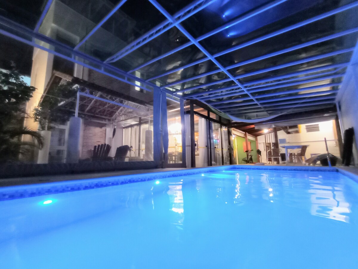 An inviting indoor pool is illuminated with soft blue lighting, surrounded by expansive glass walls. The area maintains a clear view of the outdoor space, demonstrating a blend of indoor and outdoor living. Nearby seating is visible, providing a comfortable area for relaxation.