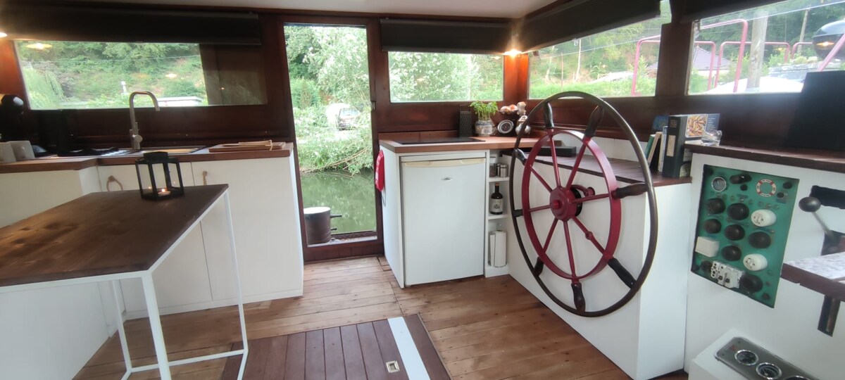 An open kitchen area features a wooden floor complemented by a white table and a robust steering wheel. Cabinets and appliances, including a fridge and a green control panel, are arranged along the walls. Natural light illuminates the space, with views of greenery through large windows.