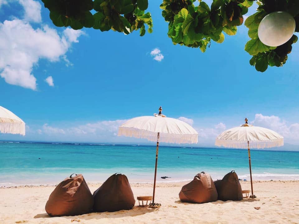 A serene beach scene is depicted, showcasing clear blue water and a sandy shore. Several large bean bags are arranged under shaded umbrellas. Green foliage frames the scene, enhancing the coastal atmosphere.