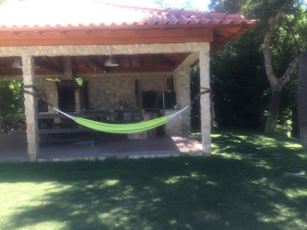 A shaded outdoor area features a stone structure with a wooden roof. A vibrant green hammock is suspended, inviting relaxation. Lush grass surrounds the space, complemented by trees in the background. A stone barbecue is also visible, enhancing the outdoor cooking experience.