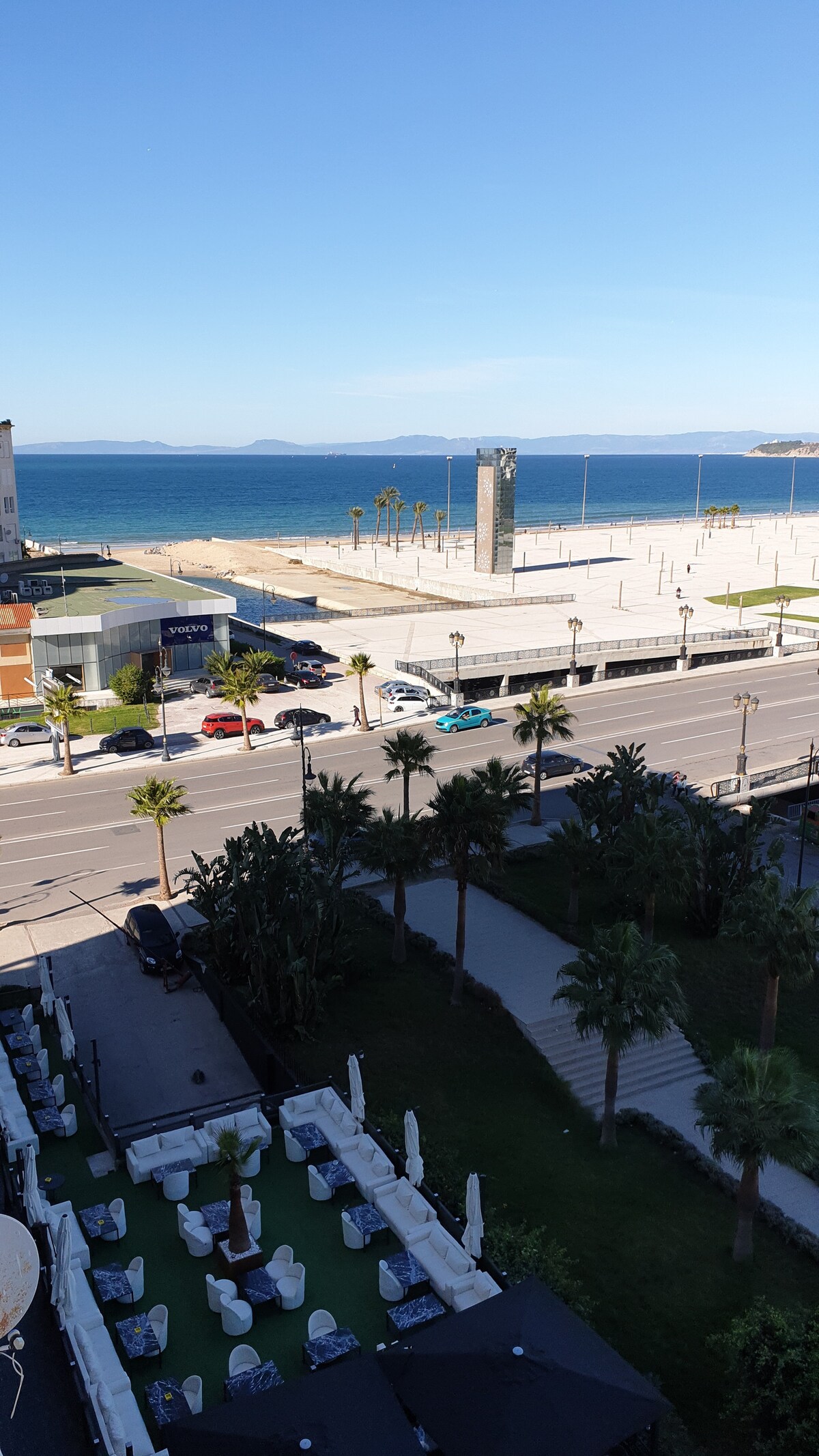 A panoramic view of the beach and ocean is captured from the apartment. The foreground features a spacious terrace with outdoor seating. A sandy beach stretches along the coastline, lined with palm trees and a clear blue sky. The area is vibrant and welcoming.