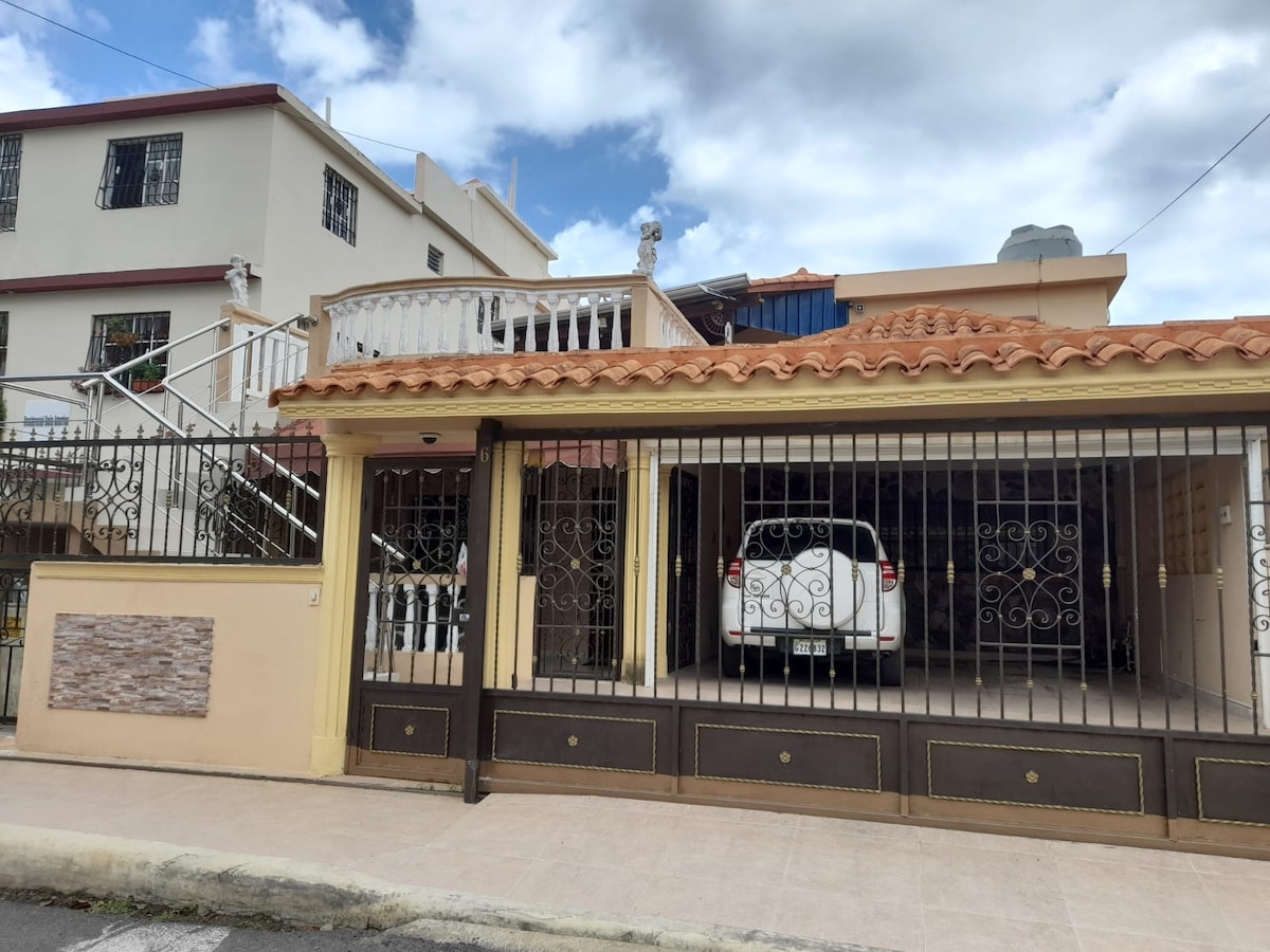 A gated entrance leads to a spacious garage displaying a parked white vehicle. The property features a two-story structure with a terracotta roof and ironwork details. A staircase can be seen ascending to a balcony. The sky above is partly cloudy.