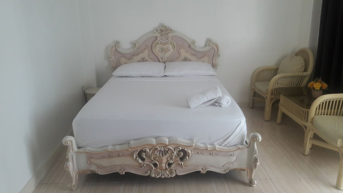A spacious queen bed is situated against a light-colored wall, featuring an ornate headboard with intricate detailing. Two comfortable chairs are positioned nearby, along with a small table. Natural light filters in through dark curtains, enhancing the inviting ambiance of the room.