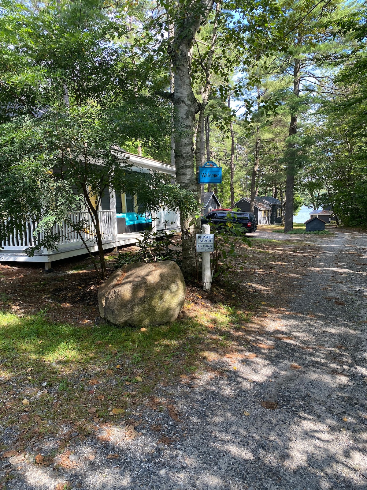 A gravel pathway leads to a cottage surrounded by tall trees. A large boulder is positioned to the side, while a sign indicates the property. The porch features blue chairs, inviting relaxation in a natural setting.