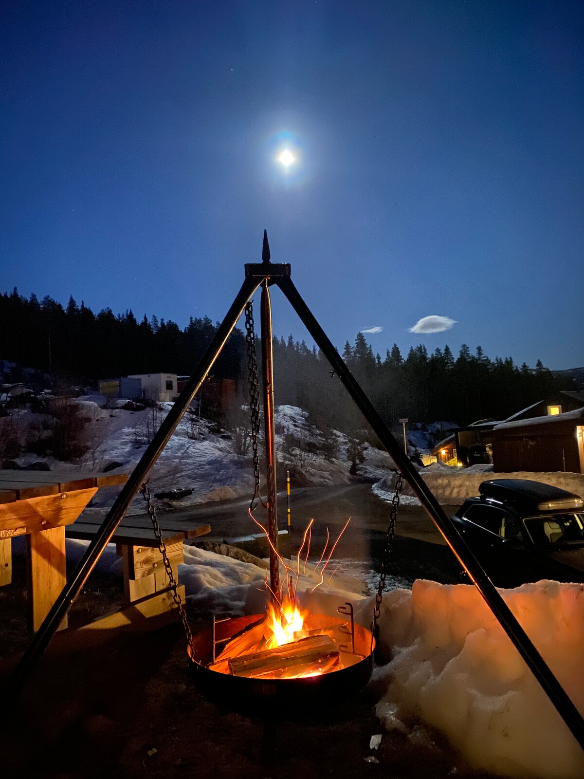 A fire pit is displayed under a clear night sky, with flames dancing above logs. A bright moon illuminates the surroundings, casting shadows on the snow-covered ground and nearby wooden structures. Trees are visible in the background, enhancing the serene atmosphere.