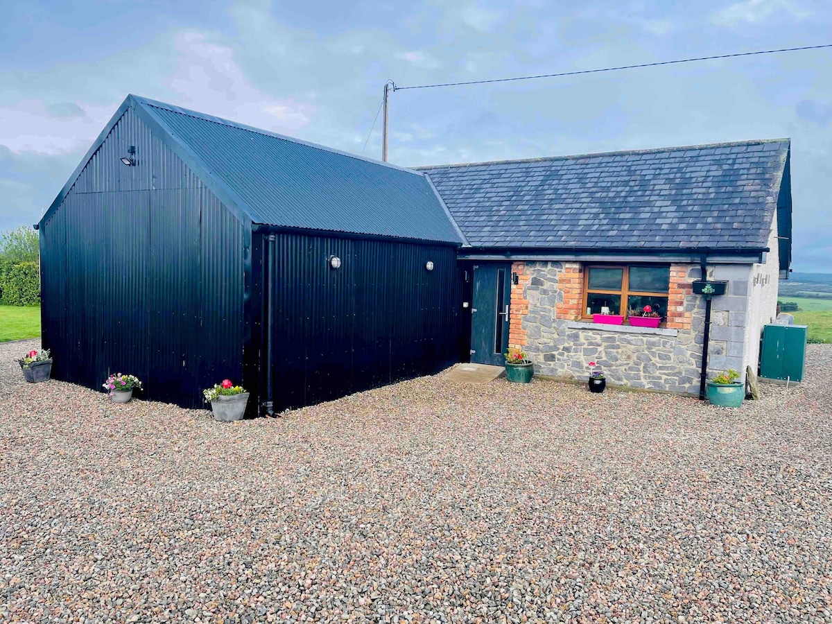 The exterior of a cottage is showcased, featuring a combination of stone and dark cladding. Colorful flowerpots line the entrance, and the overall setting is surrounded by a gravel driveway and expansive greenery in the background.