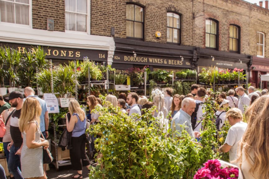 A bustling street market is filled with people browsing various stalls. Lush greenery and vibrant plants are displayed prominently, alongside a shopfront labeled 'Borough Wines & Beers.' Sunlight brightens the scene, showcasing the lively atmosphere of the market.